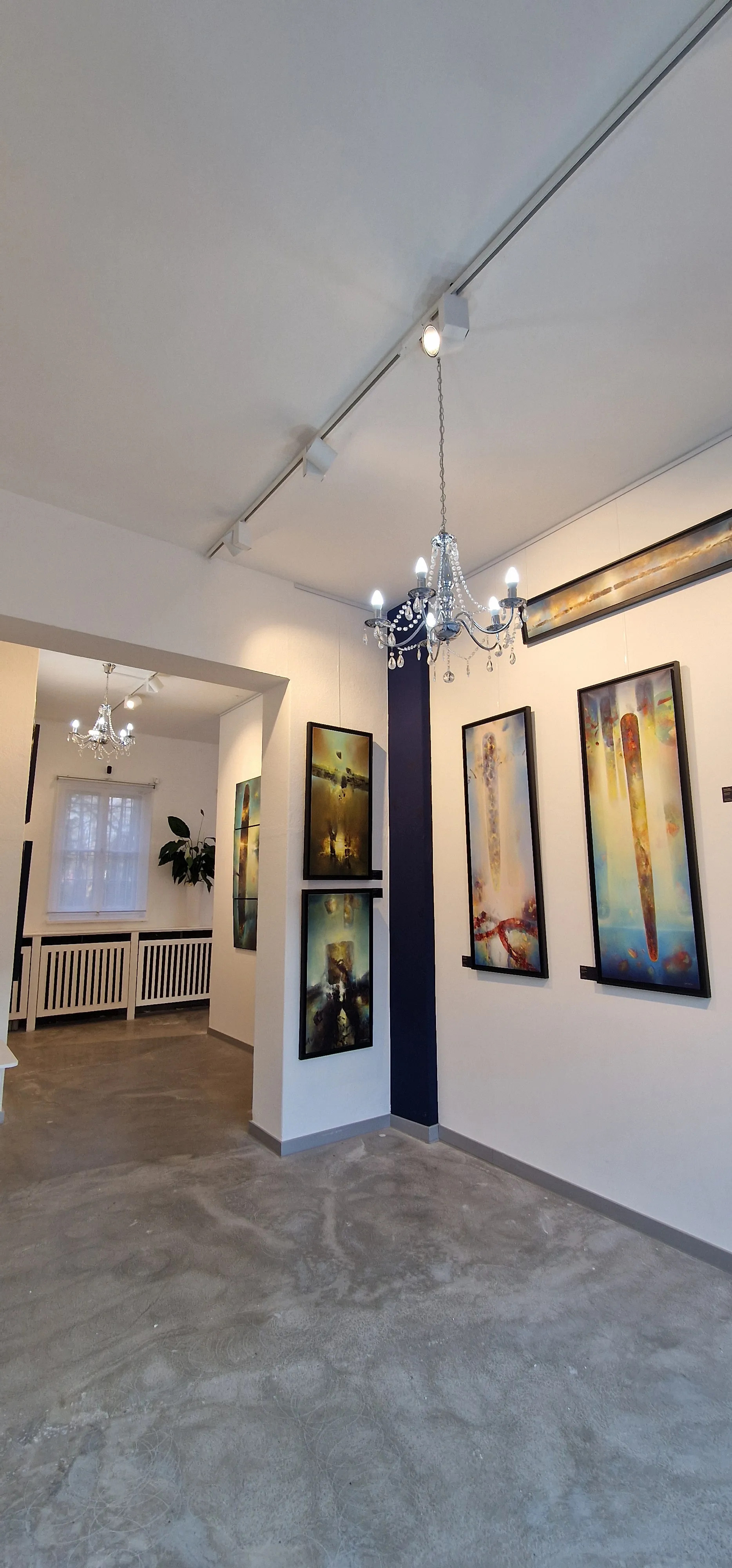Galerieflur der DNT mit hochformatigen, gerahmten Werken an weißer Wand und einem Kristallkronleuchter unter der Decke.