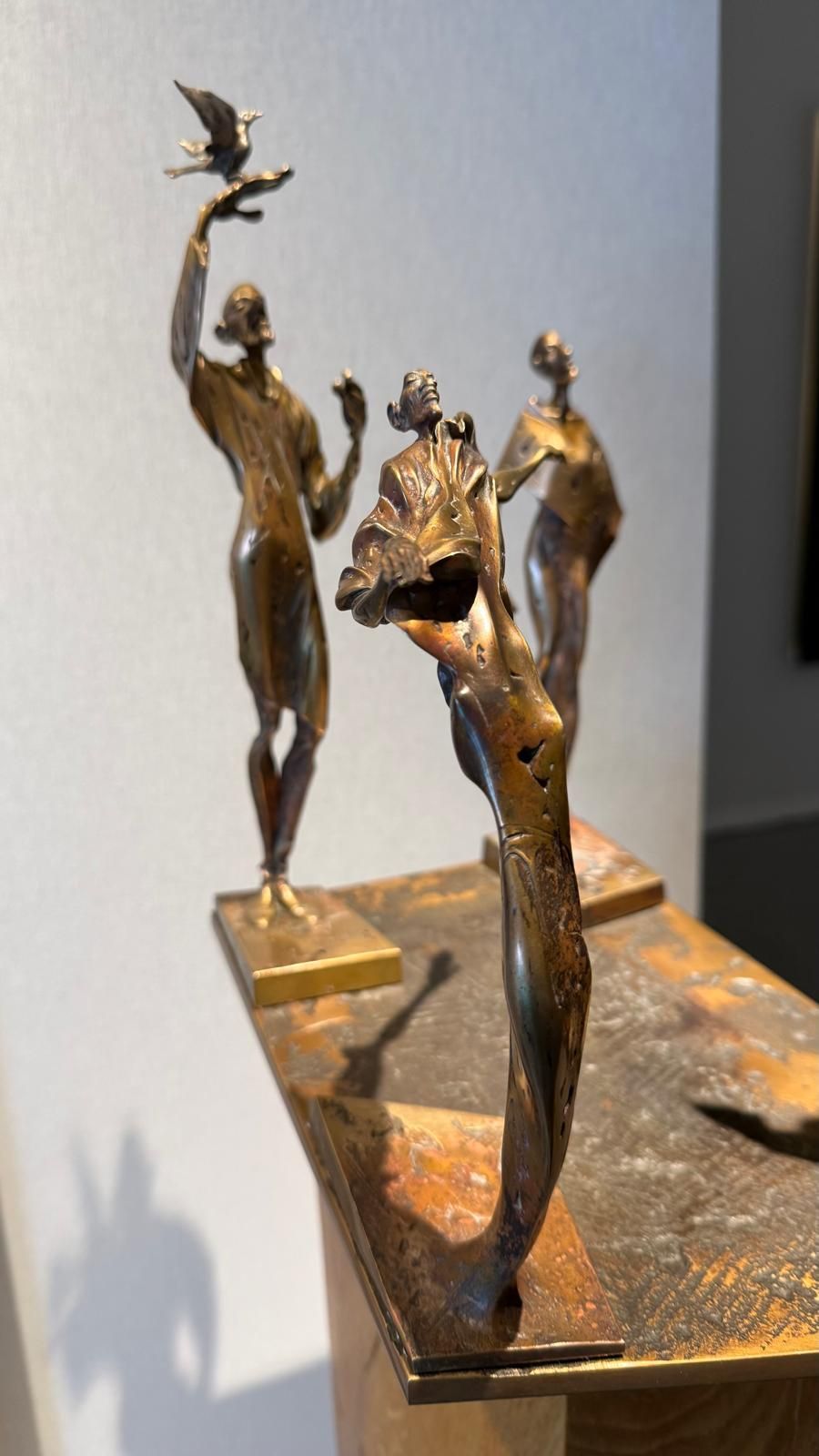 Vordere CuSi3 Siliziumbronze-Skulptur „Herbst" (ca. 25 cm): schlanke Figur in starker Rückbiegung mit anliegendem Gewanddetail, patinierte Oberfläche in warmen Bronze- und Brauntönen, auf Metallplatte vor hellgrauer Galeriewand; zwei weitere Figuren unscharf im Hintergrund.